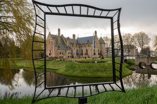 View of Radboud Castle framed by a decorative metal sculpture, overlooking a calm moat with green grassy banks and willow trees in Medemblik, Noord-Holland, Netherlands.