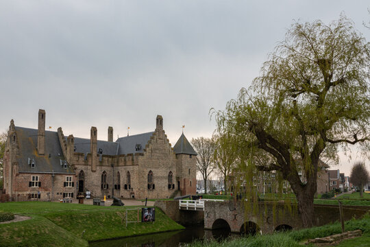 View of Radboud Castle with its medieval brick walls, a stone bridge over the moat, and a large willow tree under a cloudy sky in Medemblik, Noord-Holland, Netherlands.