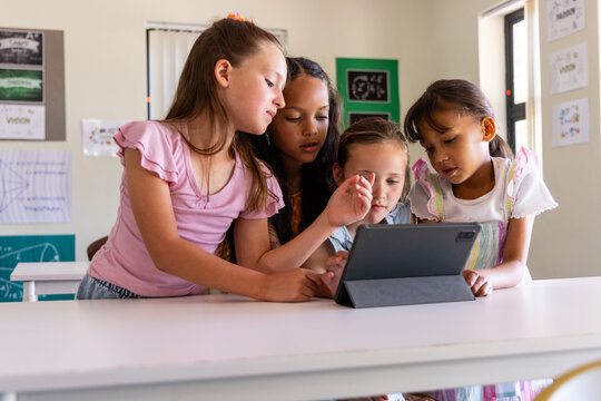 Diverse female elementary students leaning, pointing at tablet with folio on white classroom table