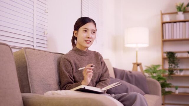 young Asian women writing journal notebook sitting sofa relaxing home living room planning daily schedule creative ideas diary personal thoughts lifestyle