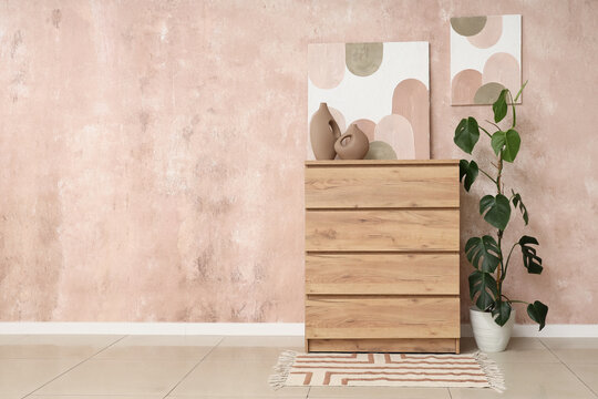Wooden commode with vases, palm plant and paintings on pink grunge wall in room