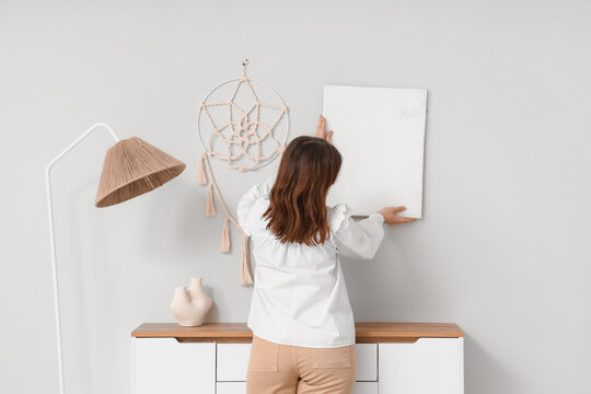 Young woman hanging painting above commode on light wall in room, back view