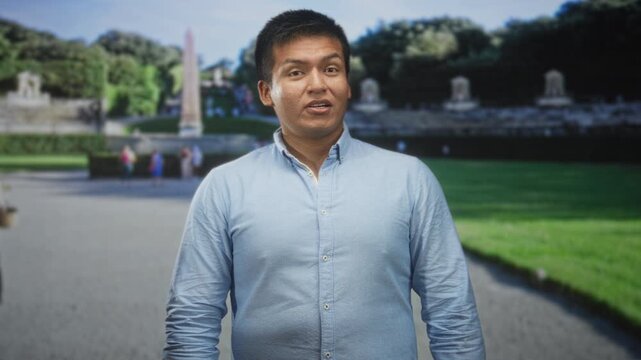 Man, young hispanic, in a light blue button shirt looking to the side with hands relaxed on street by a stone obelisk and fountain; calm contemplation.