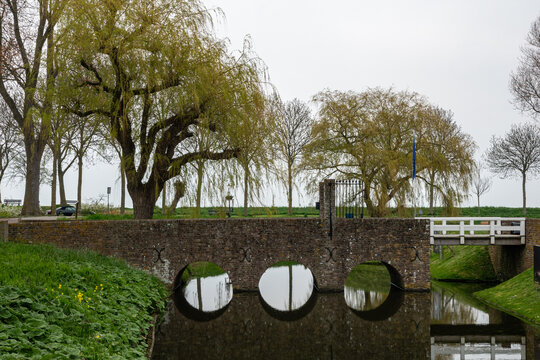 View of the historic stone bridge at Radboud Castle with circular arches reflecting in the moat water and weeping willow trees in Medemblik, Noord-Holland, Netherlands.