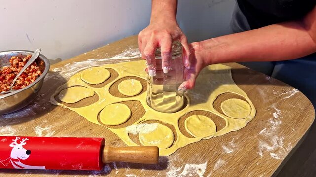 Hands cutting dough circles with glass for dumplings on floured kitchen table