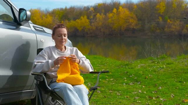 Woman knitting outdoors during an autumn car trip. Middle-aged woman enjoying her hobby, sitting in a folding chair next to her car and knitting a yellow scarf while appreciating the autumn by river