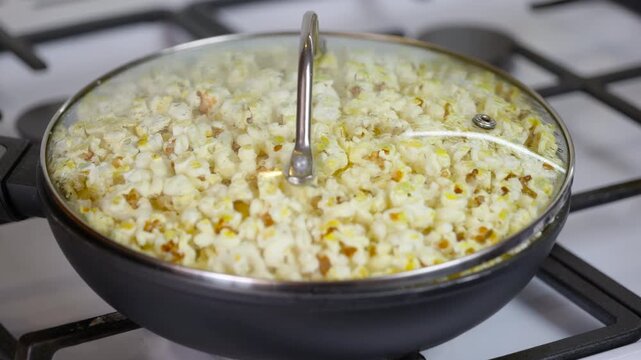 Popcorn popping in a pan on gas stove cooking snack. Popcorn kernels popping inside a hot pan with a glass lid on a gas stove, creating a delicious and popular cinema snack for movie nights