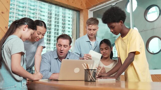 Caucasian teacher giving comment or advise student while smart bot pointing at laptop screen at classroom. Diverse student taking a note while teacher teach lesson while using computer. Edification