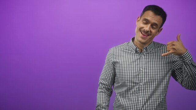 Young hispanic man with moustache wearing a checked shirt makes a call me hand gesture and points with his finger in studio; playful.