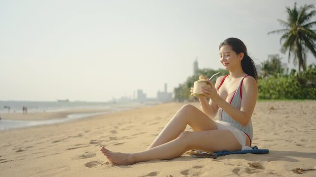 Beautiful young Asian woman in a swimsuit relaxes on a sandy beach, putting down her smartphone to enjoy the tropical ocean breeze, sunshine, and sipping fresh coconut water during her vacation.