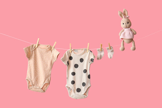 Baby bodysuits with socks and toy hanging on rope against pink background