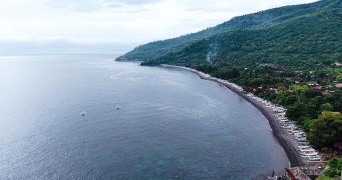 Aerial panorama of Amed Beach in Bali, Indonesia, showing traditional fishing boats lined along the volcanic shoreline with calm ocean waters and lush tropical hills in the background.