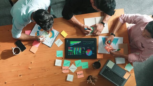 Top aerial view of diverse team writing colorful sticky notes to brainstorm marketing idea while looking at financial chart. Multicultural smart business people taking a note about plan. Convocation.
