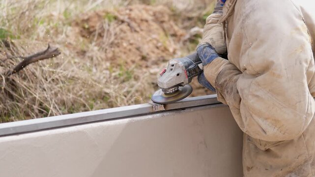Worker Uses Angle Grinder On Rail, Closeup Of Gloved Hands Smoothing Metal Edge With Abrasive Disc, Sparks And Dust Fly, Outdoor Fence Construction Site, Dusty Jacket And Protective Gloves, Methodical