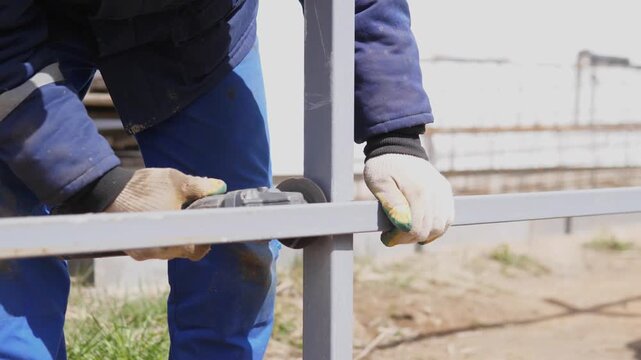 Closeup Worker Aligning Steel Rail, Gloved Hands Steadying Horizontal Bar Outdoors, Using Measuring Tool And Clamp, Dirt Ground, Focused Manual Labor, Precise Alignment For Fence Installation