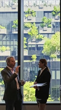 Two professionals talk by a large window overlooking a modern green building with terraces of plants, blending business with sustainable urban design.