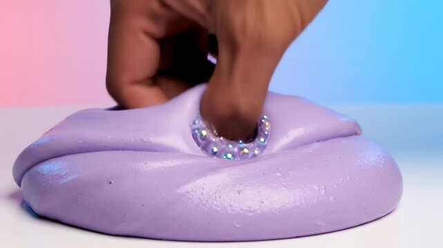 Person's hand pressing, stretching, and adding shiny rhinestones to a satisfyingly smooth and vibrant purple slime on a white surface with a colorful pink and blue background. asmr