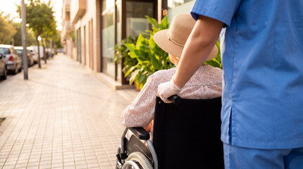 Healthcare professional in gloves assists a senior woman in a wheelchair on a sunny street, providing mobility. © Stephen
