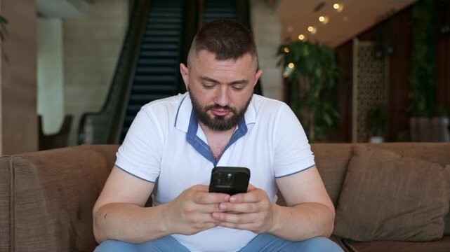 Man with a beard dressed in a white polo shirt and light blue pants sits on a couch, focused on his smartphone in a contemporary indoor space featuring escalators and plants. Footage