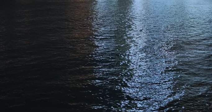 Top static view, dark water sparkles in light at night. Calm water surface of river, lake at night. Abstract background with blue, orange light reflections from nearby city. Tranquil, mysterious mood