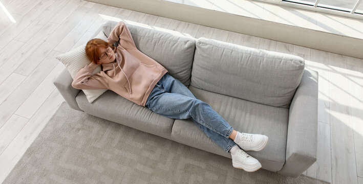 Redhead teenage girl lying on sofa near large window at home, top view