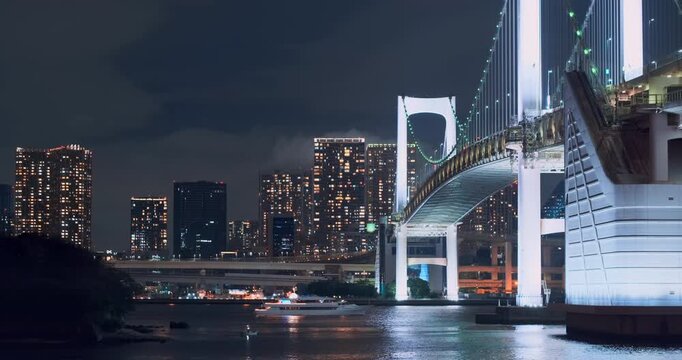 Luminous modern Tokyo metropolis at night, static wide view. Famous rainbow bridge over bay. Shining city skyscrapers is reflected in river. boat cruising on water under dark, cloudy sky in Japan