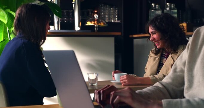 Slow Motion Of Women Talking Over Coffee With Laptop In Modern Caf&eacute;. Two women discussing ideas while sharing coffee at a caf&eacute; table with a laptop, representing remote work and collaboration.