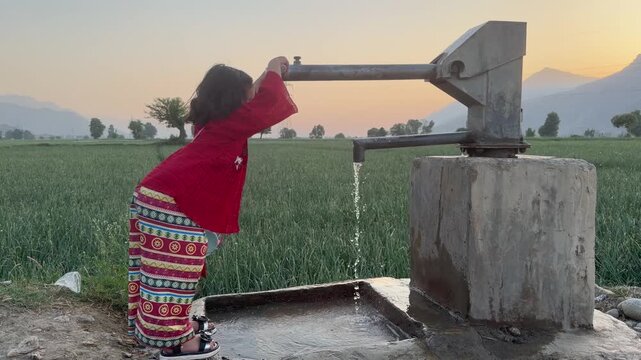 A young girl in traditional clothing uses a manual hand pump to draw fresh water in a rural setting.