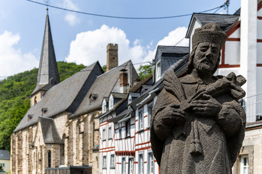 View of the stone statue of Saint John of Nepomuk with the historic Dreifaltigkeitskirche church and traditional half-timbered houses under a blue sky in Monreal, Rhineland-Palatinate, Germany.