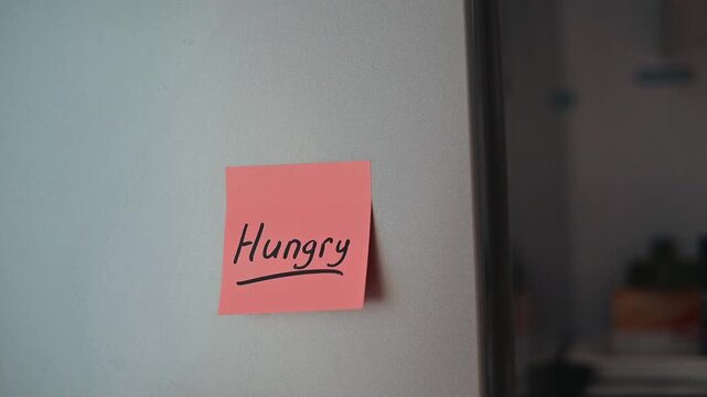 A bright kitchen shows a sticky note with the word hungry written on it. The note is placed on a fridge door, creating a simple and relatable moment about food needs