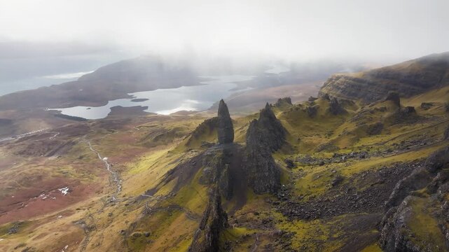Slow cinematic drone pan over the Storr rock formation, Isle of Skye, Scotland, thick blanket of cloud overhead, mist pooling in the valley and rising up the cliff face