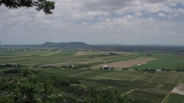 A high-angle landscape view features a vast expanse of agricultural fields and orchards stretching toward distant hills under a cloudy sky, framed by trees in the foreground