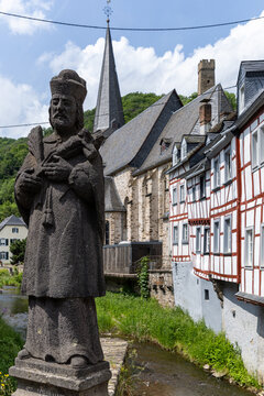View of the stone statue of St. John of Nepomuk by the Elzbach river with half-timbered houses and the parish church in Monreal, Rhineland-Palatinate, Germany.