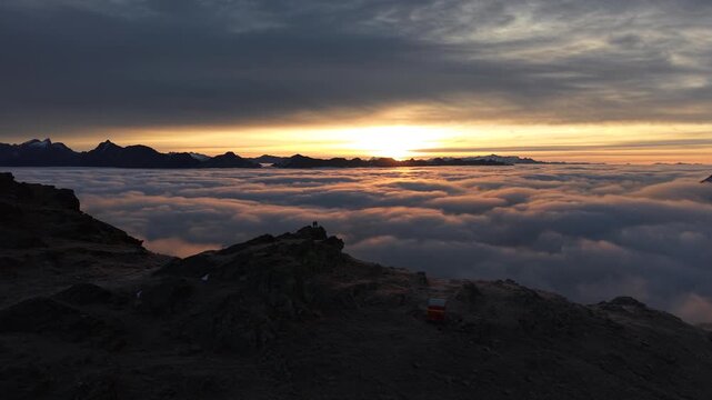 Aerial flyover above hikers enjoying the sunrise glow over the inversion of clouds in the Swiss Alps with a panoramic view of the mountains and colorful sky