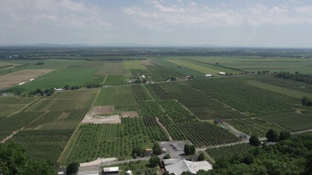 Wide aerial view of vast agricultural farmland and apple orchards under a partly cloudy sky, featuring organized rectangular fields, farm buildings, and distant mountain ranges