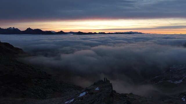 Aerial flyover over hikers enjoying the sunrise glow over the inversion of clouds in the Swiss Alps with an alpine panoramic view of the mountains and colorful sky at dawn