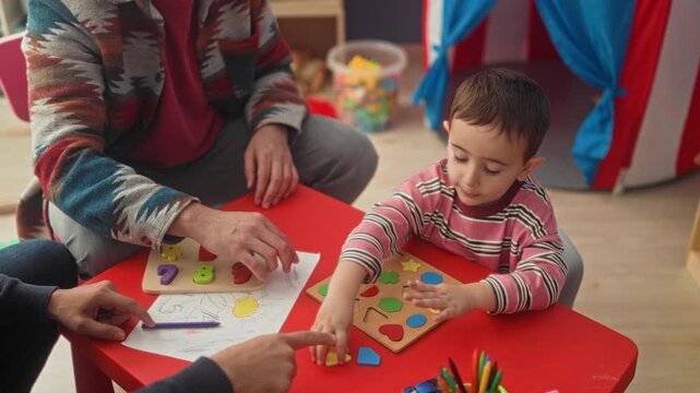 Boy placing wooden shapes into a puzzle on a red table in a preschool playroom, adult hands guiding and child raising hand for a high five; learning joy.