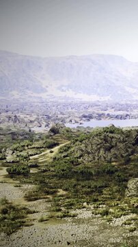 A rocky landscape stretches out, with patches of green plants scattered among the stones. Distant mountains rise under a clear sky. The scene shows natural beauty and variety in terrain.