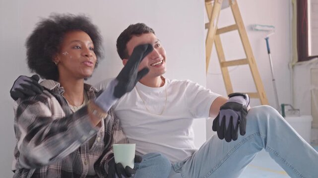A young couple shares a joyful moment during their home renovation project. The woman, with her curly hair, and the man, both wearing gloves, sit together, laughing and enjoying a break.
