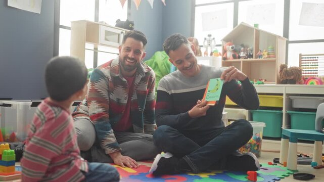 Boy kneels on foam playmat and shows an orange card to teacher while seated in a preschool building; gentle learning joy.