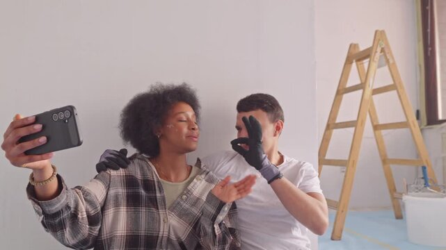 A diverse couple, the woman with curly hair and the man, both smiling, take a selfie in a bright, modern room with a wooden ladder in the background.