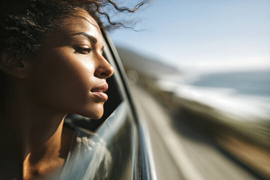 Young Black woman driver leaning from car window with eyes closed, sunlit curly hair blowing and side profile against blurred coastal road