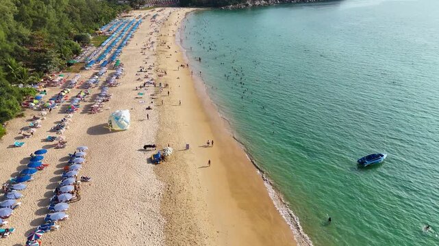 Aerial view of sunny tropical beach full of people on vacation with colorful parasols and turquoise ocean water in Phuket, Thailand.