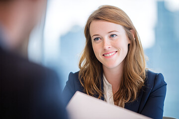 Fototapeta premium Smiling redheaded female lawyer in navy blazer and white blouse reviewing documents while listening attentively in bright high - rise office