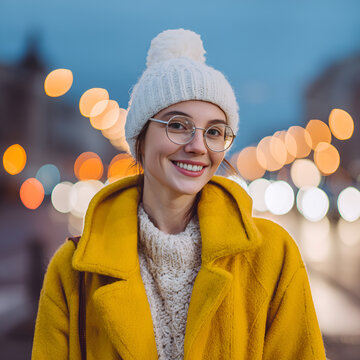 Uśmiechnięta kobieta w białej czapce i ż&oacute;łtym płaszczu na miejskiej ulicy &ndash; zimowa moda, okulary i wieczorne światła w tle.