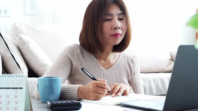 Smiling Asian woman having an online meeting on a laptop from home. Concept of remote work, video conference, e-learning, and professional business communication in a comfortable living room office