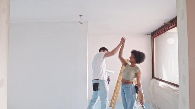 A young couple collaborates on a home renovation project, their hands joined as they step across a freshly painted wall. The room, bathed in natural light from a large window.