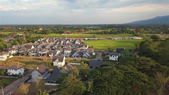 Cinematic aerial 270 orbit over a quiet eastern community Features vibrant rooftops and green farming plots toward distant mountain peaks during a soft sunset in a wide regional territory