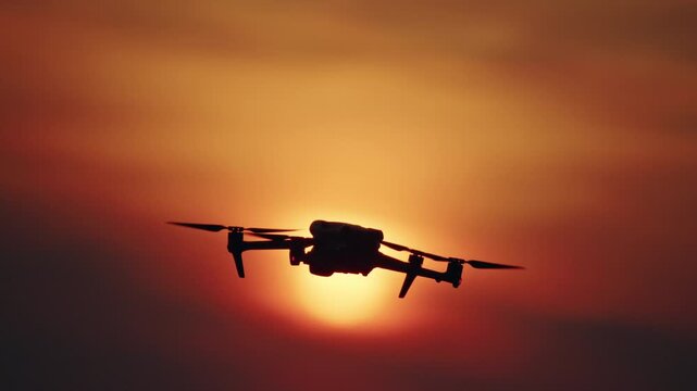 Drone silhouette flying across glowing sunset sun with orange sky