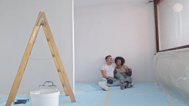 A couple shares a joyful moment in a sunlit, partially renovated room. The man, casually dressed in a white t-shirt and jeans, sits on a protective blue sheeting on the floor.